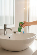 Pack Caffè-Dipendente — 4 dentifrici ÉclatDent: un tubetto tenuto sopra un lavabo bianco, bagno luminoso e rubinetto cromato