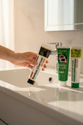 Pack Splendore Carbone — 4 dentifrici ÉclatDent: un tubetto tenuto sopra un lavabo bianco, bagno luminoso e rubinetto cromato